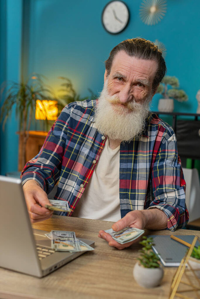 Mutlu kıdemli adam para sayıyor ve merkez ofiste dizüstü bilgisayar kullanıyor. Başarılı serbest çalışan büyükbaba ofisten bir tomar para alıyor kar paylarını hesaplıyor maaş gelirlerini. Dikey - Fotoğraf, Görsel