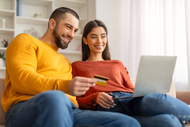 Kolay ödemeler. Laptop bilgisayarı ve kredi kartı olan mutlu genç çift evdeki koltukta dinleniyor, neşeli eşleri online alışveriş yapıyor, İnternet alışverişlerinin tadını çıkarıyor, - Fotoğraf, Görsel