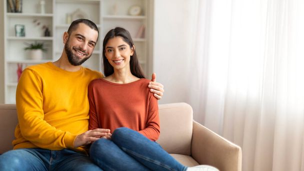 Ajánlat pároknak. Szerető férfi és nő ül szorosan a kanapén és mosolyog a kamera előtt, romantikus házastársak ölelkeznek, miközben pózolnak a modern nappali belső terében, felidézve az otthon és a kényelem érzését - Fotó, kép