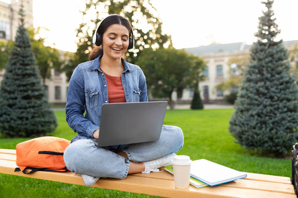 Молодая индийская студентка имеет онлайн-урок, сидя на скамейке в университетском городке, используя ноутбук и беспроводные наушники, копировальное пространство. Электронное обучение, современное образование - Фото, изображение