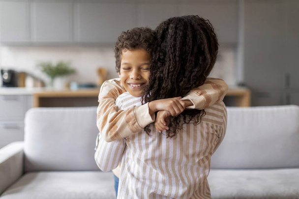 Vreugdevolle zwarte tiener jongen met heldere glimlach omarmen zijn aanhankelijke moeder in gezellige, goed verlichte woonkamer, gelukkig Afrikaans Amerikaans gezin van twee delen tedere moment, zoon knuffelen moeder en glimlachen - Foto, afbeelding