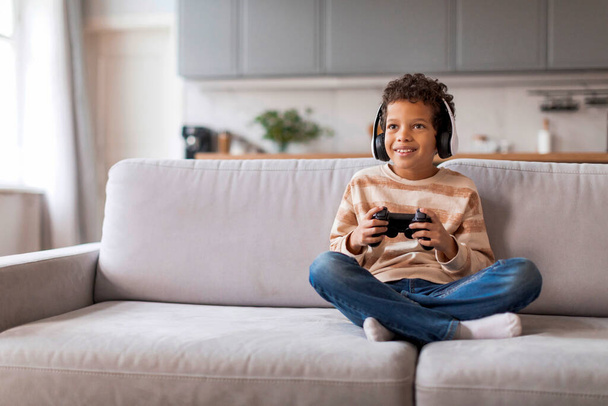 Cheerful black boy wearing wireless headphones playing with game controller, happy african american male kid holding joystick, enjoying video games while sitting on couch in living room - Photo, Image