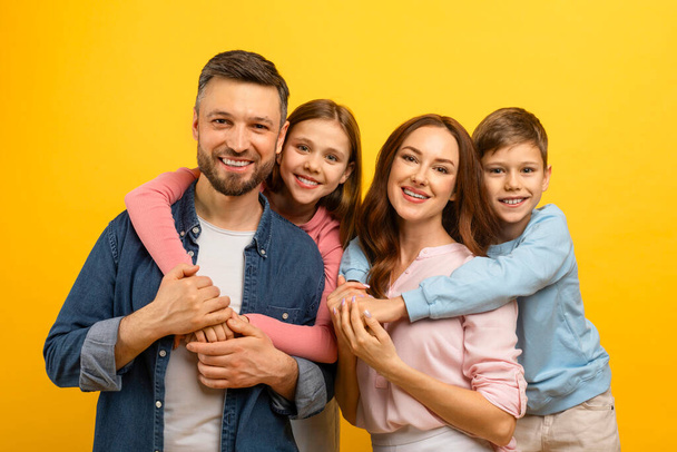Cheerful caucasian family of four posing together against yellow background, little son and daughter hugging happy young parents, sharing joyful moment on bright studio backdrop, free space - Photo, Image
