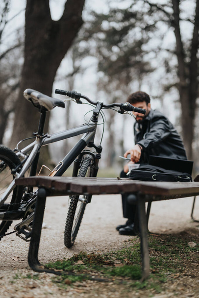 Un uomo d'affari bello ed elegante si concentra sul suo computer portatile mentre lavora all'aperto in un parco urbano sereno, con la sua bicicletta accanto a lui. - Foto, immagini