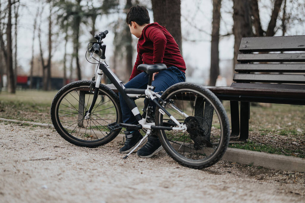 Nuori yksilö lepää puistonpenkillä polkupyörä vieressään nauttien rauhallisesta hetkestä ulkona.. - Valokuva, kuva