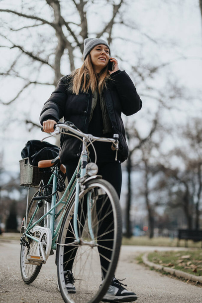 Radostná žena v neformálním oblečení a beanie se usmívá během telefonního hovoru při jízdě na kole na stezce v klidném prostředí parku, ilustrující multitasking a aktivní životní styl. - Fotografie, Obrázek