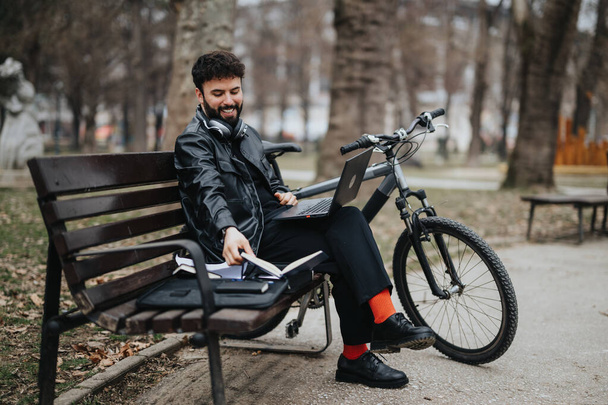 Stylowy mężczyzna przedsiębiorca wielozadaniowy zdalnie z laptopem i notatnikiem w parku miejskim, wraz z rowerem. - Zdjęcie, obraz