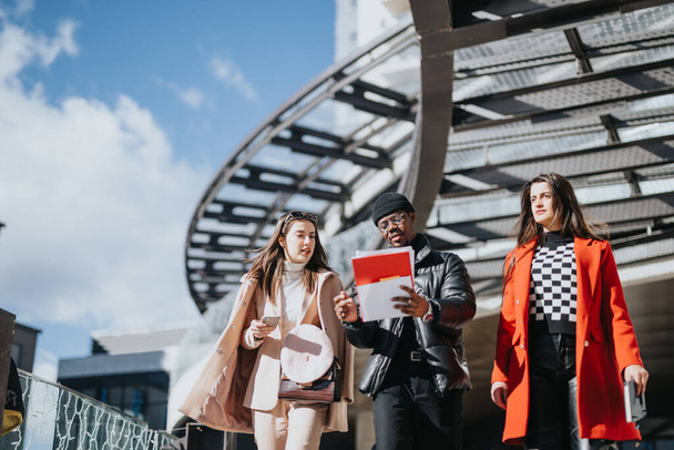 Young business colleagues in stylish outfits walk together outside, engaged in discussion with a digital tablet in hand under modern architecture. - Photo, Image