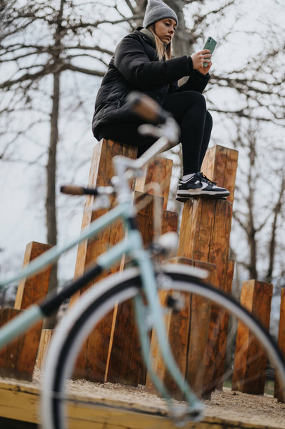Uma mulher casualmente vestida sentada em troncos de madeira em um parque enquanto olha para o telefone. Uma bicicleta embaçada em primeiro plano adiciona uma sensação de lazer e atividade ao ar livre. - Foto, Imagem