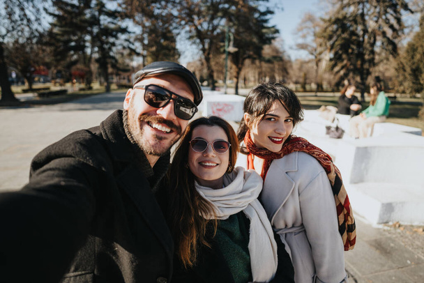Tři šťastní přátelé různých etnik sdílející radostný okamžik, kdy se skupinka selfie v slunečný zimní den ve veřejném parku. - Fotografie, Obrázek