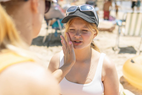 На фоне летнего пляжа, мать нежно наносит солнцезащитный крем на лицо дочери, когда они сидят в купальниках, воплощая суть отдыха на море и родительского ухода... - Фото, изображение