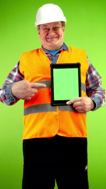 Старший будівельник у жилеті та шоломі, що показує планшет з порожнім рекламним дисплеєм. Зрілий працівник тримає пристрій на зеленому екрані з копіювальним простором. Зелений екран. Ключ хроми. Вертикальний. - Кадри, відео