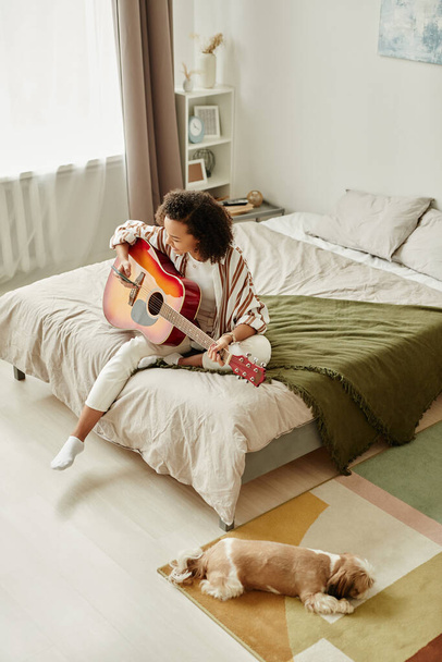 Luftiges Hochwinkel-Porträt einer jungen Frau, die im Bett Akustikgitarre spielt, während sie es sich zu Hause mit Hund gemütlich macht - Foto, Bild