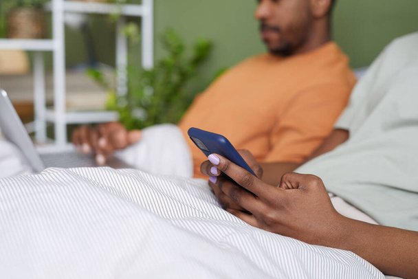 バックグラウンドで彼女のボーイフレンドとベッドに横たわっている間,スマートフォンにメッセージを入力する若い女性のクローズアップ - 写真・画像