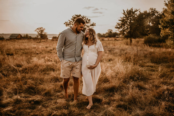 Happy family pregnant caucasian blonde woman with moles on face in white cotton dress walks with husband meadow summer. man in light natural clothes and shorts holds hand wife. trees in sunbeams - Photo, Image