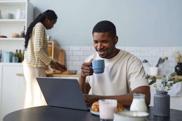 Afro-Amerikalı bir adam mutfakta kahvaltı yaparken dizüstü bilgisayar kullanıyor. Karısı da arkada yemek pişiriyor. - Fotoğraf, Görsel