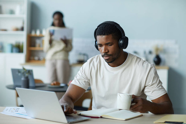 Afrikkalainen amerikkalainen mies langattomissa kuulokkeissa juo kahvia ja työskentelee verkossa kannettavan tietokoneen kotona - Valokuva, kuva