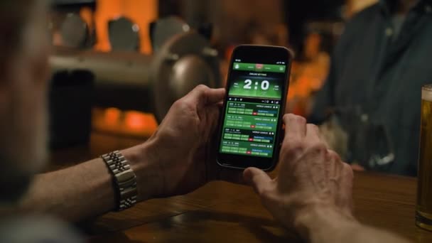 Close up of mature sports fan checking game score and bookmaker ratings in app using mobile phone. Caucasian man bets on football match online sitting at bar counter in pub at night. Gambling concept. - Footage, Video