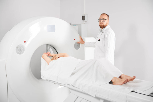 Medical computed tomography or MRI scanner. Male radiologist presses MRI button to examine female patient, looking to the camera. - Photo, Image