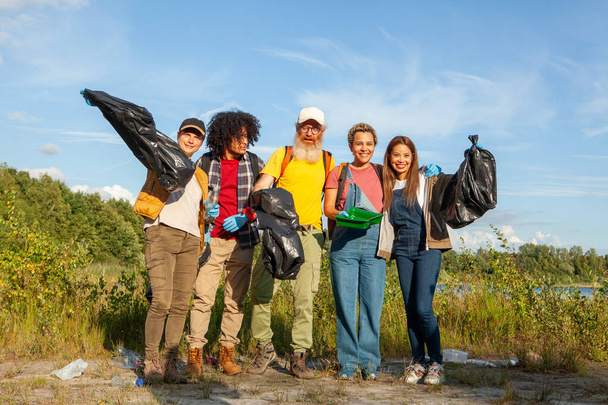 Μια ομάδα πέντε διαφορετικών εθελοντών στέκεται περήφανα με τα συλλεχθέντα απορρίμματα, επιδεικνύοντας την περιβαλλοντική τους διαχείριση. Αφιερωμένο Εθελοντές Επιδρώντας Θετικά στο Περιβάλλον με τη Λίμνη Καθαρισμού. Υψηλή - Φωτογραφία, εικόνα