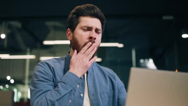 Sleepy businessman yawning and covering mouth with hand while sitting at desktop in modern office. Bored man with tired eyes working on laptop and looking at screen while dreaming about weekends. - Footage, Video