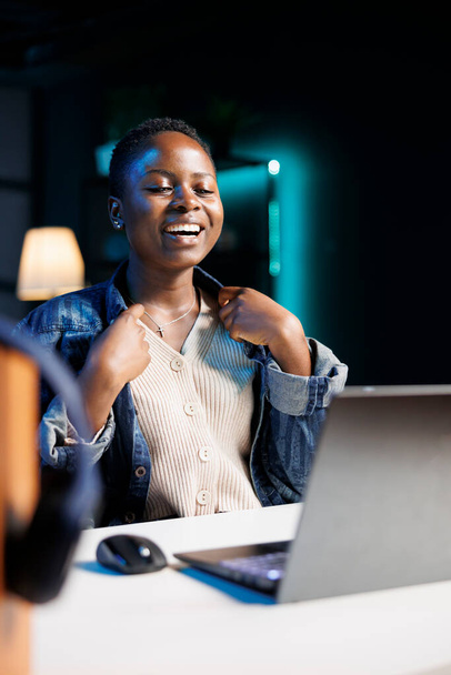 Czarna kobieta uśmiecha się szeroko i szczęśliwie podczas korzystania z cyfrowego komputera osobistego w swoim mieszkaniu. Podekscytowany amerykański bloger siedzący przed laptopem, mający wideokonferencję telefoniczną. - Zdjęcie, obraz