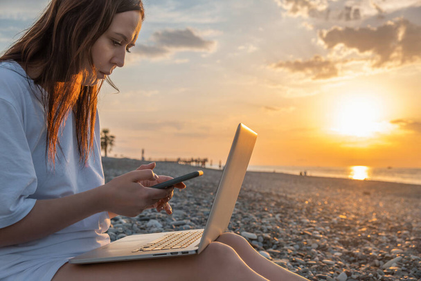Digitaalinen nomadinainen työskentelee kannettavan tietokoneen ja matkapuhelimen kanssa rannalla auringonlaskun aikana. Nainen freelancer työtä verkossa älypuhelimella ja tietokoneella etäisyydeltä. Etätyö lomalla - Valokuva, kuva