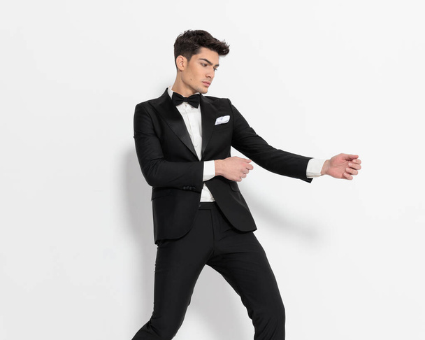 крутой элегантный бизнесмен в черном туфле держит руки в стороны, смотрит в сторону и модно позирует на сером фоне  - Фото, изображение