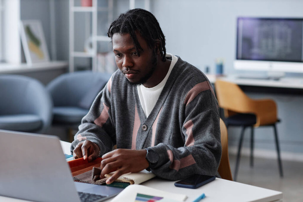 Afro-Amerikaanse jonge ontwerper werkt op laptop en met monsters van stof zitten op zijn werkplek in kantoor - Foto, afbeelding