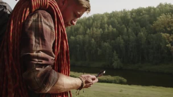 Arc shot του νεαρού άνδρα τουρίστα με σχοινί αναρρίχησης και σακίδιο πλάτης στέκεται στο λόφο, χρησιμοποιώντας πυξίδα και κοιτάζοντας την κατεύθυνση, ενώ πεζοπορία στη φύση για τις καλοκαιρινές διακοπές - Πλάνα, βίντεο