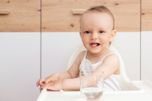 Süßes kleines Mädchen mit Glas und Trinkwasser. Glückliches Baby im Babystuhl in der Küche. Kind trinkt Wasser. - Foto, Bild
