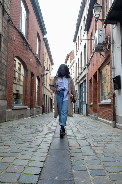 Dit beeld toont een stijlvolle vrouw die door een geplaveide straat loopt in een historische Europese wijk. Ze is modieus gekleed in een blauw hemd met kraag, hoge taille jeans, een beige trench coat - Foto, afbeelding