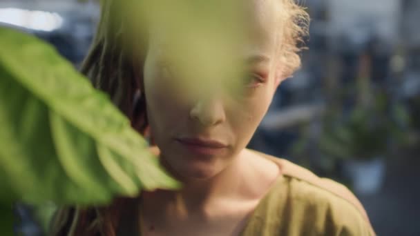 Середній крупним планом портрет молодої кавказької жінки з неперевершеним поглядом позують на камеру серед зелених листя рослини в приміщенні - Кадри, відео