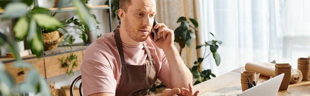 Un hombre en una tienda de plantas, sentado en una mesa, ocupado en una llamada telefónica. - Foto, imagen