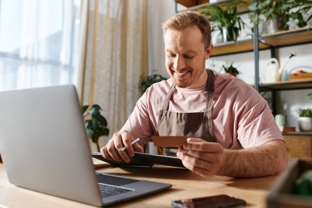 Ein Mann sitzt an einem Laptop, hält eine Kreditkarte in der Hand und kauft vermutlich Nachschub für sein kleines Geschäft.. - Foto, Bild