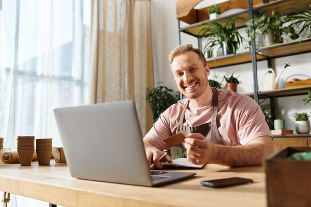Um homem com um laptop e cartão de crédito, focado em seu pequeno negócio em uma loja de plantas. - Foto, Imagem