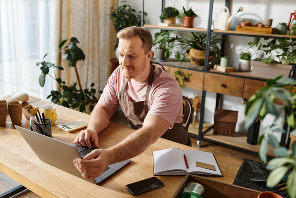 Egy divatos ízlésű férfi ül az asztalnál, a laptopjára koncentrál egy buja növénytömb közepette egy otthonos növényboltban.. - Fotó, kép