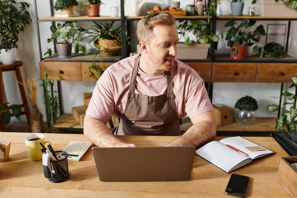 Ein Mann konzentriert sich auf seinen Laptop an einem Schreibtisch, umgeben von Pflanzen in einem Pflanzengeschäft Einstellung. - Foto, Bild