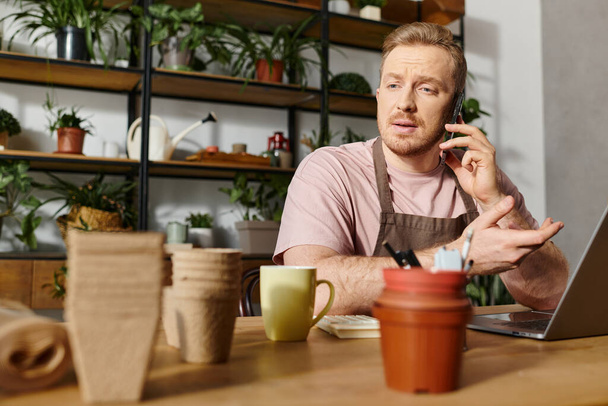 Ein Mann sitzt an einem Tisch in einem Pflanzenladen und telefoniert. Das Ambiente verströmt eine Atmosphäre der Kleinunternehmer. - Foto, Bild