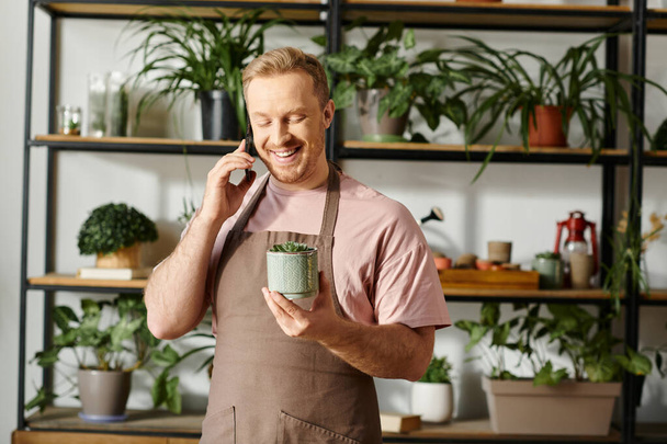 Чоловік у фартусі багатозадачний, тримаючи рослинний горщик і розмовляючи на мобільному телефоні в ботанічному магазині. - Фото, зображення