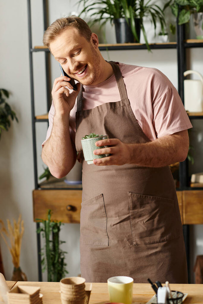 Un hombre guapo en un delantal multitarea mientras conversa en un teléfono celular en una tienda de plantas. - Foto, imagen