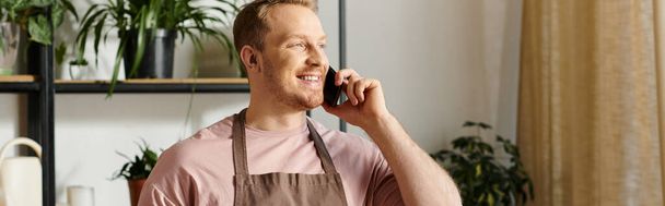 Ένας κομψός άντρας με ποδιά συνομιλεί σε ένα κινητό τηλέφωνο, διευθύνει το εργοστάσιό του με έμφαση στην εξυπηρέτηση πελατών.. - Φωτογραφία, εικόνα