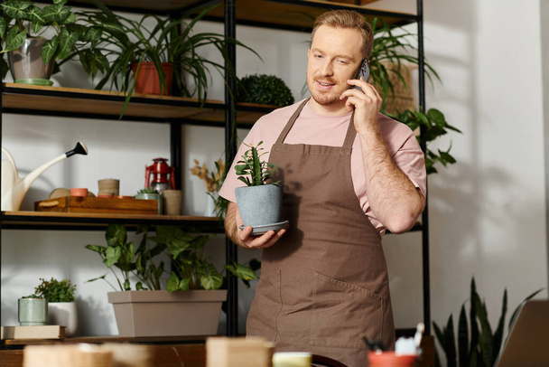 Un hombre multitarea, sosteniendo una planta en maceta y hablando en un teléfono celular en una tienda de plantas. - Foto, imagen