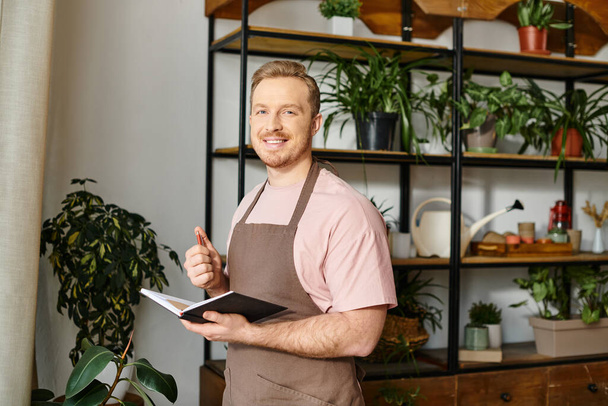 Un hombre guapo en un delantal sostiene un cuaderno, manejando su tienda de plantas con una dedicación de dueños de negocios. - Foto, imagen