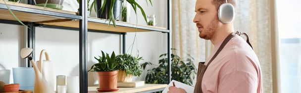 Um homem elegante vestindo fones de ouvido fica na frente de uma prateleira em uma loja de plantas, cercado por vegetação exuberante e beleza botânica. - Foto, Imagem