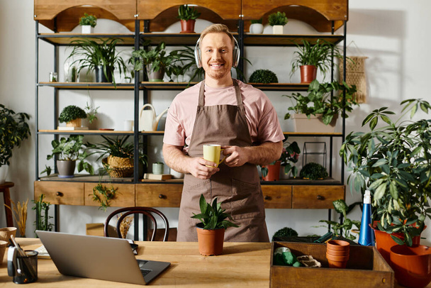 Un bell'uomo con un grembiule che tiene una tazza davanti a un portatile in un negozio di piante. - Foto, immagini