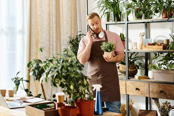 Um homem em um avental fala em um telefone celular enquanto mantém uma planta em vaso em uma loja de plantas. - Foto, Imagem