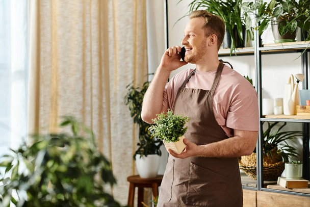 Un homme bavardant sur un téléphone portable et tenant une plante en pot dans un magasin de plantes, incarnant le multitâche et la propriété de l'entreprise. - Photo, image