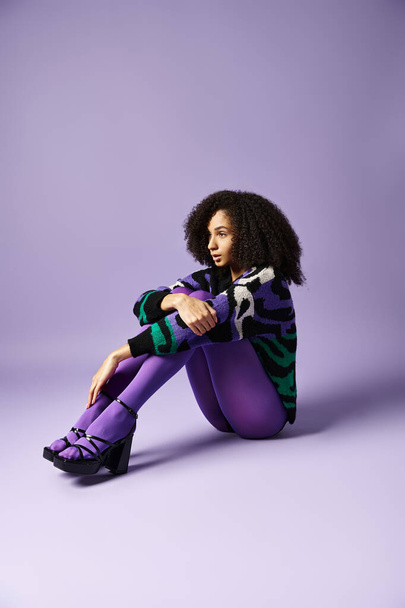 Молода жінка в яскравих колготках і светрі сидить витончено на землі на фіолетовому фоні. - Фото, зображення