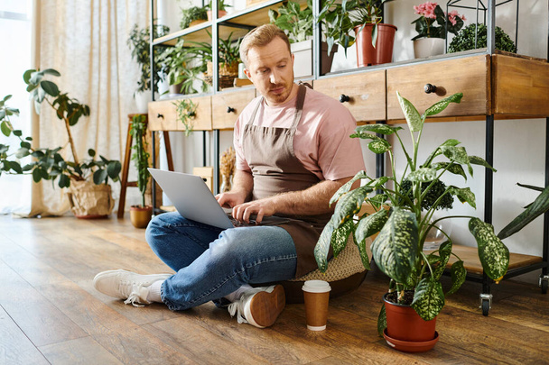 Ein Mann in einem Pflanzenladen sitzt auf dem Boden und arbeitet angestrengt an seinem Laptop und verkörpert die Hingabe an sein kleines Geschäft. - Foto, Bild